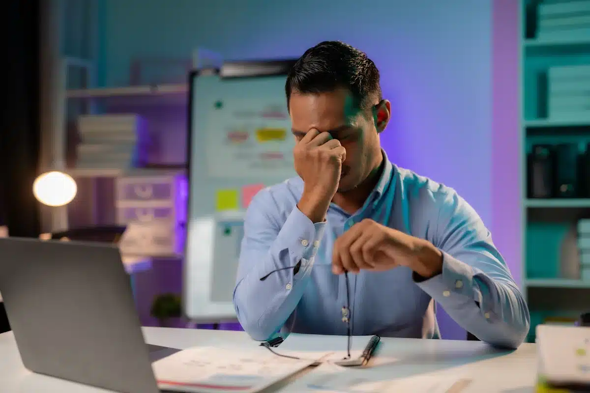 A remote or hybrid male worker suffering from burnout sitting at his desk in front of his laptop rubbing his eyes with his eyeglasses in his hand