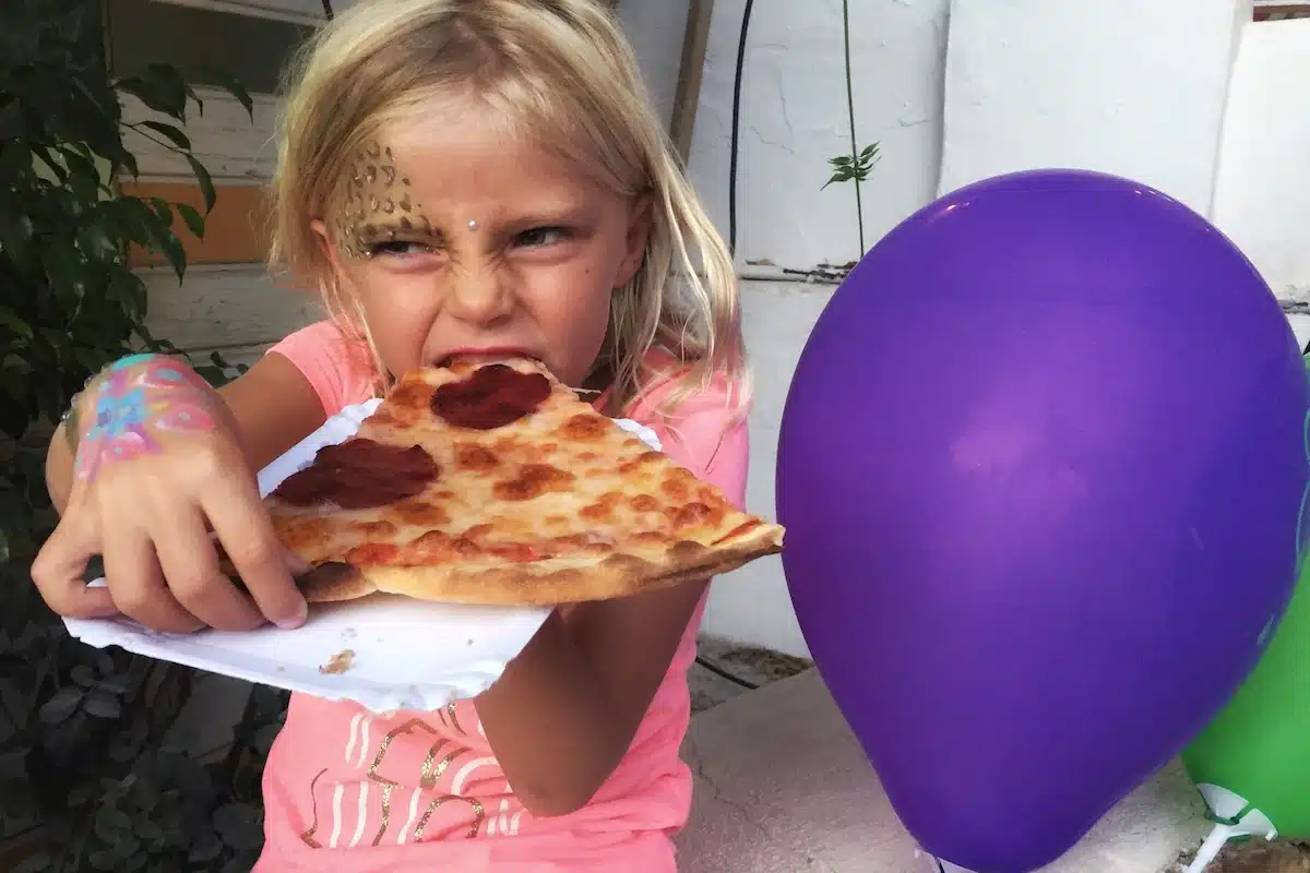 A young neice enjoying a slice of pizza with a purple ballon to her side