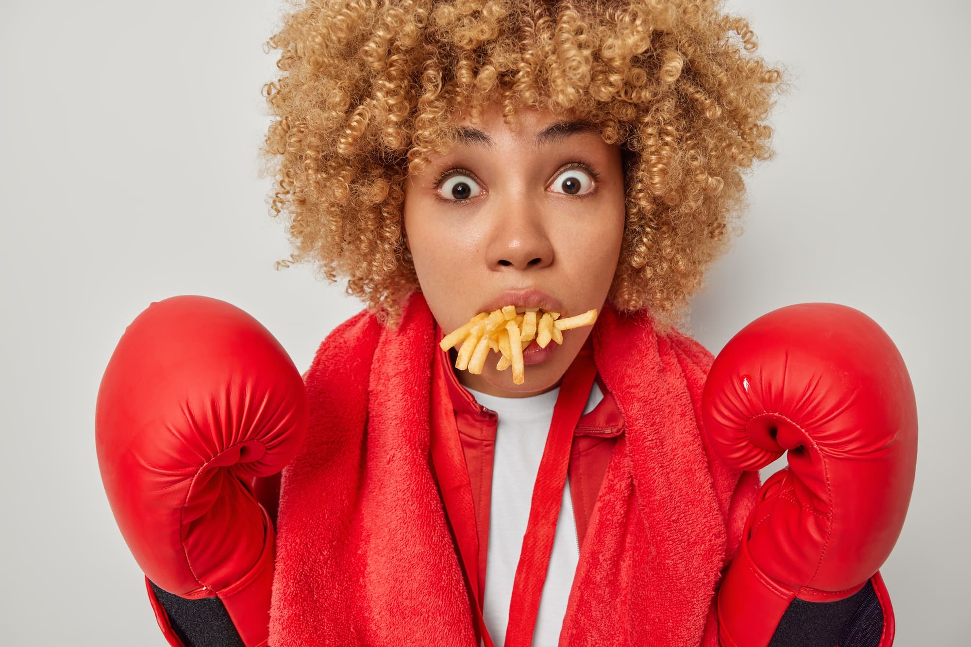 curly-haired woman wearing boxing gloves has a mouth full of french fries. enjoys a power lunch