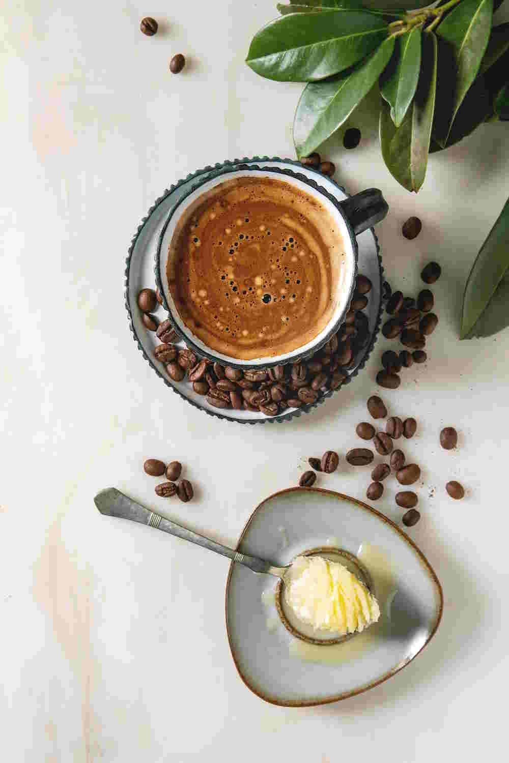 Bulletproof Coffee | Still Worth The Hype In 2026? 1 Bulletproof coffee. Keto diet coffee in blue ceramic cup with organic ghee butter in spoon with beans and green branch over white marble background.