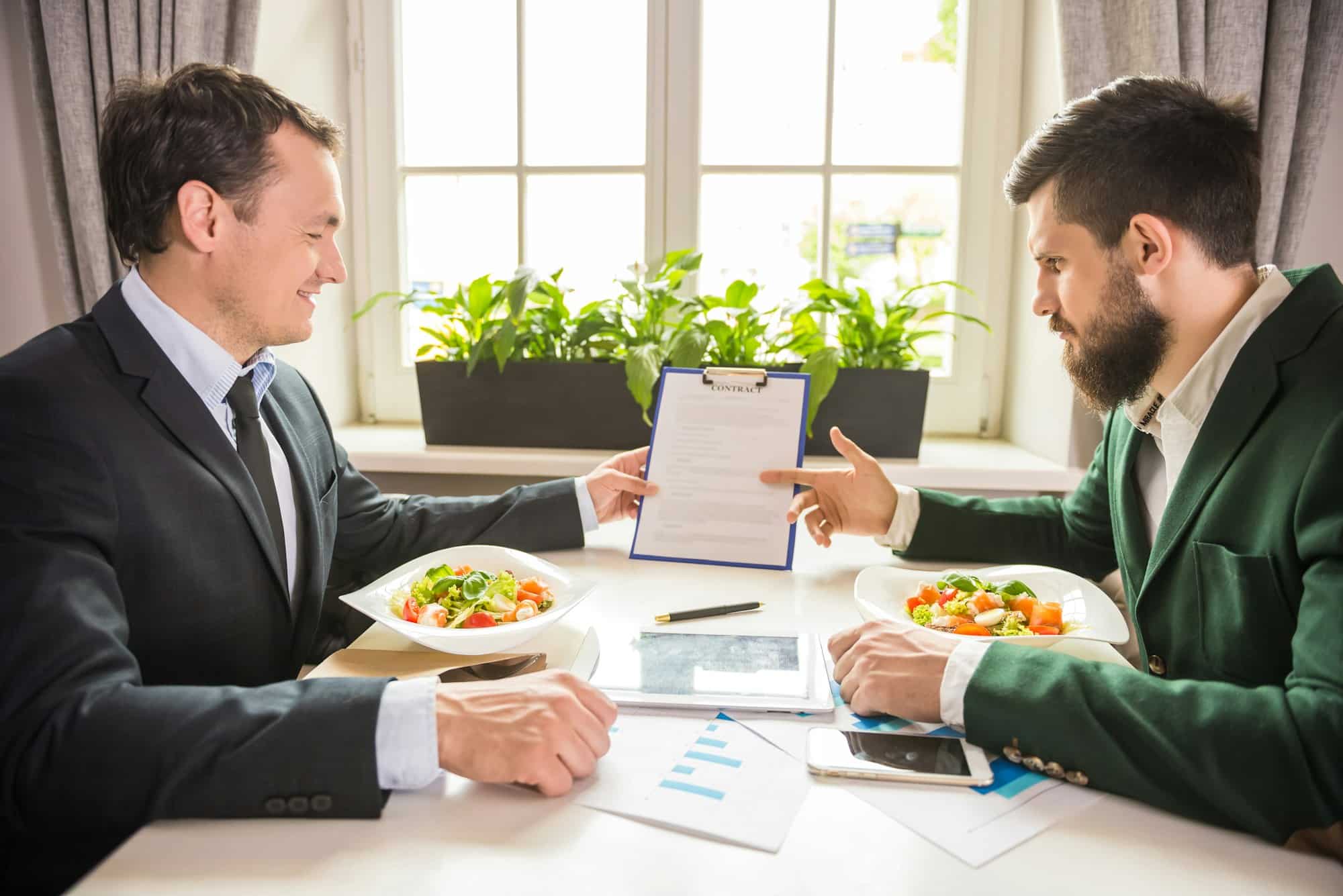 Two men seated at the restaurant when every forkful matters during a business lunch dressed in suit and tie with their salads in front of them while one is presenting a document to the other