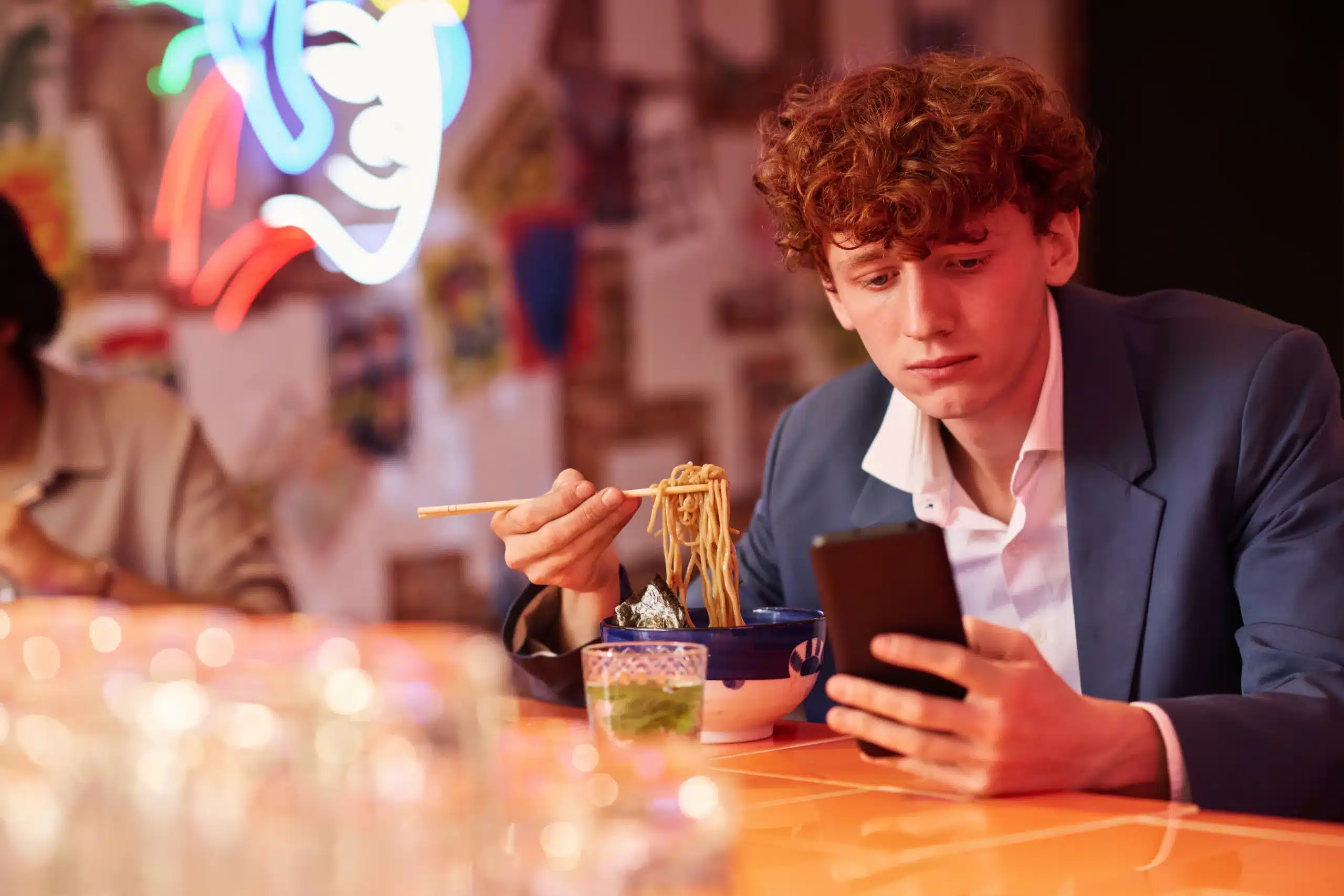 A man eating Asian noodles from a bowl while looking at his phone