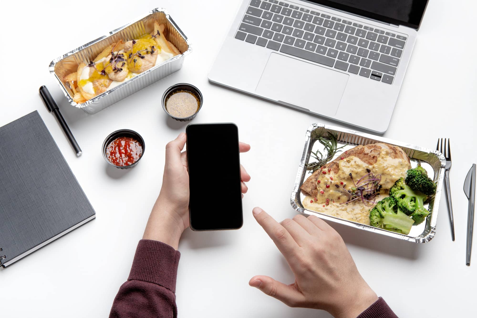 Photo of a man holding a cell phone with two aluminum containers of food on either side and a Macbook at the top of the photo. 