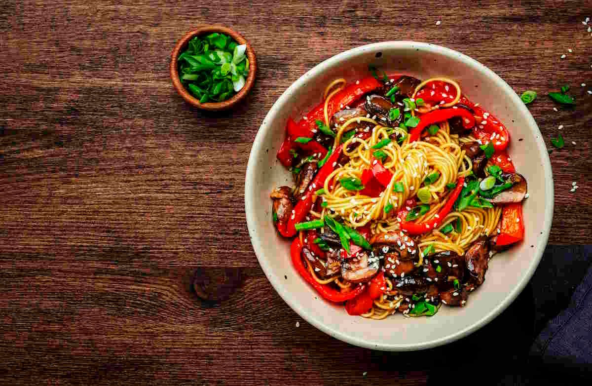 Vegetable Lo Mein Vegan Stir fry noodles with vegetables, paprika, mushrooms, chives and sesame seeds in bowl. Wooden table background, top view, copy space