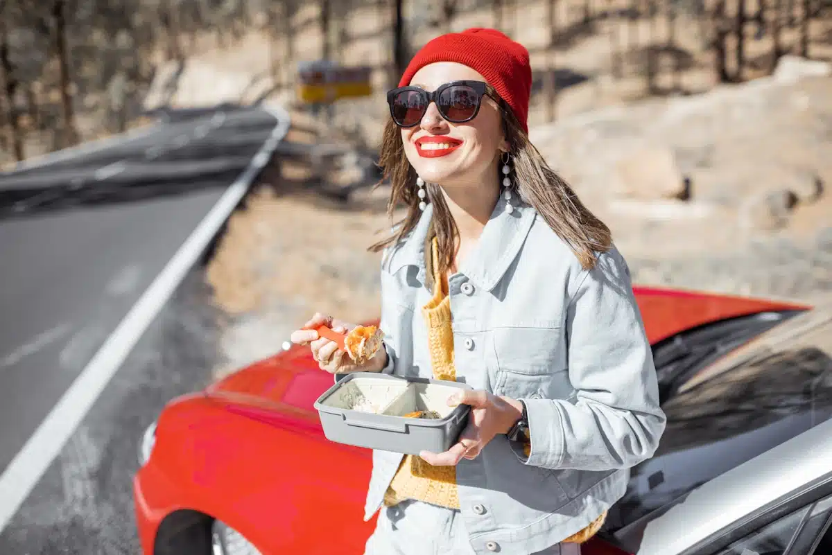 A woman eating her lunch whilew leaning on her car during a road trip in the mountains.